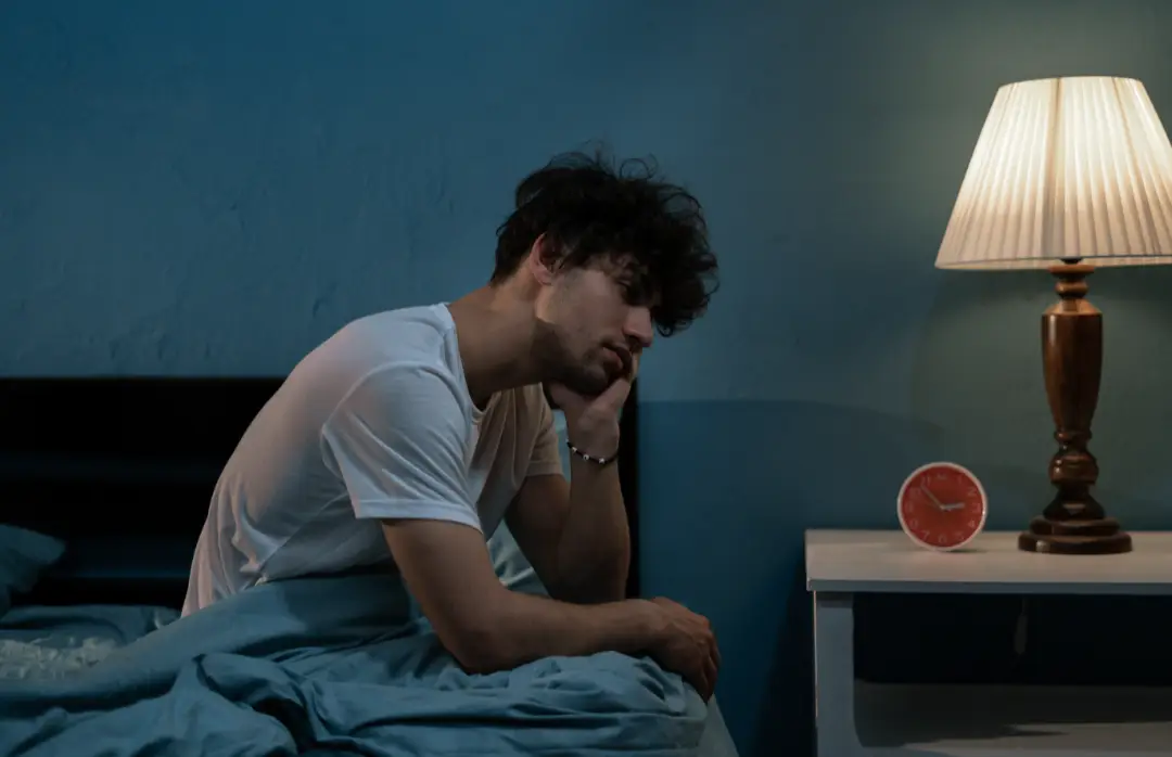 Muž sedící na posteli v nočním prostředí, vypadající ospale a unaveně, s hlavou opřenou o ruku. Vedle něj je noční stolek s rozsvícenou lampou a červeným budíkem. Pokoji dominuje tmavě modrá barva.